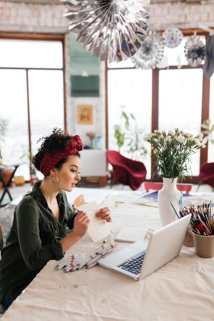 Young gorgeous woman with dark curly hair sitting at the table thoughtfully showing sketch in laptop while spending time in modern cozy workshop with big windows