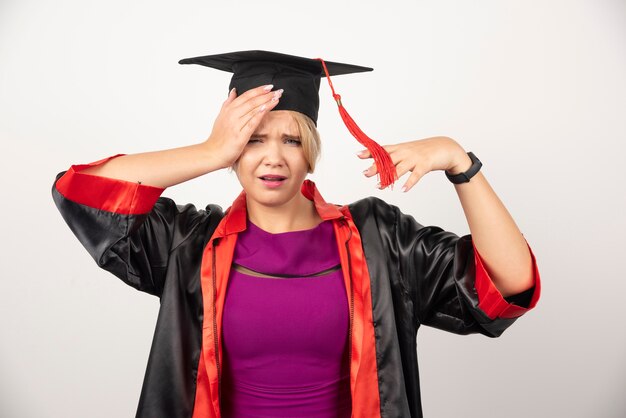 Young graduate student in gown having headache on white wall. 