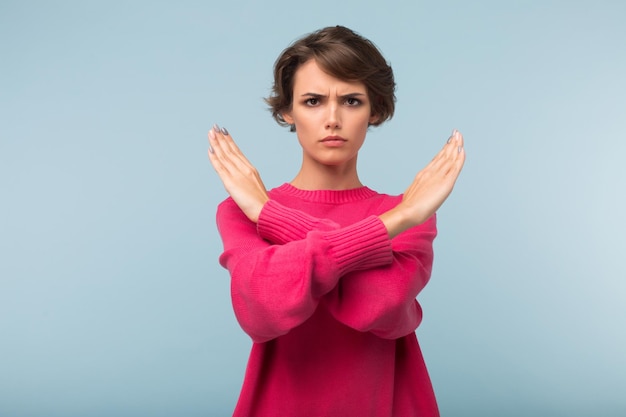 Foto gratuita giovane donna scontrosa con capelli corti scuri in maglione rosa che mostra un gesto di arresto mentre guarda con rabbia nella fotocamera su sfondo blu