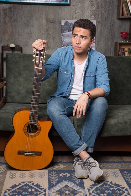 Young guitar player holding a beautiful guitar and sitting on sofa. High quality photo