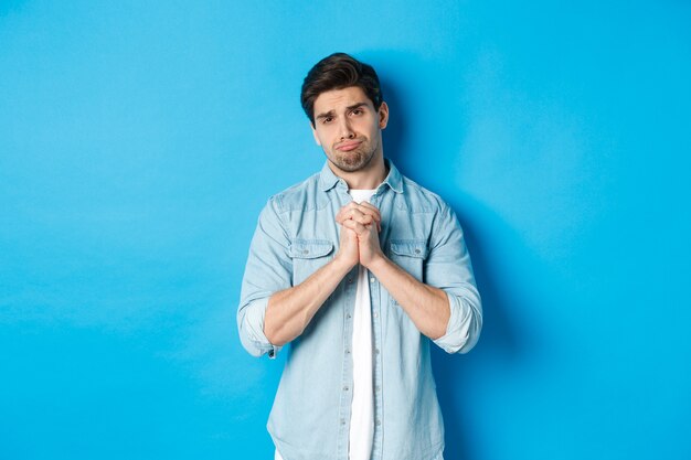 Young guy begging for help, pouting and looking sad while asking for favour, need something, standing against blue background