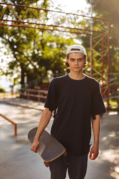 Giovane ragazzo in maglietta nera e berretto bianco che guarda pensieroso nella fotocamera mentre tiene in mano lo skateboard con lo skatepark sullo sfondo