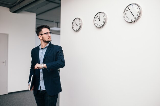 Young guy in glasses is  walking in office. He wears blue shirt, dark jacket, jeans and beard. He holds  laptop in hand. He is looking to clock on wall.