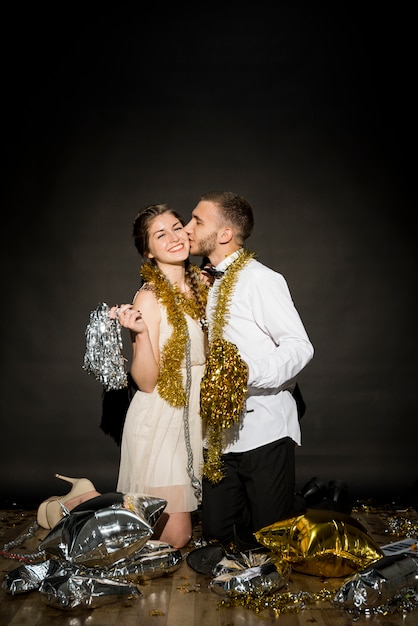 Free photo young guy kissing smiling lady in evening wear with tinsel on floor