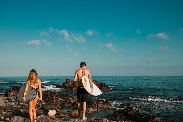 Young guy and lady with surf boards going on stone shore to water