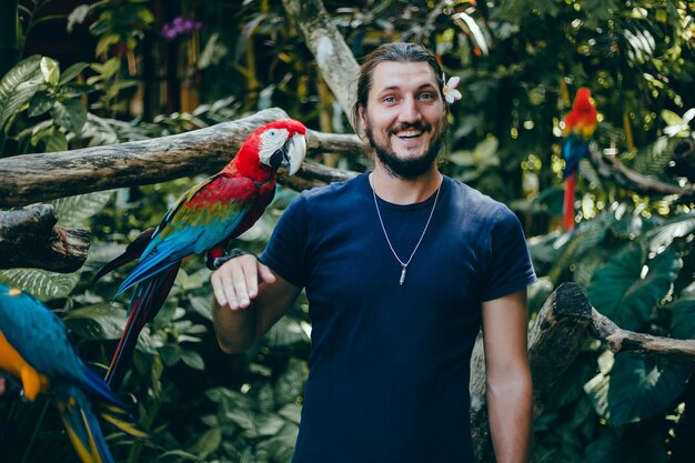 молодой парень позирует в зоопарке с попугаем в руке, бородатым мужчиной и птицей