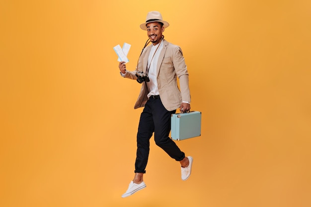 양복과 모자를 쓴 젊은 남자가 여행가방과 주황색 배경의 티켓을 들고 점프합니다. 베이지색 재킷을 입은 행복한 남자와 복고풍 카메라로 포즈를 취하는 흰색 티
