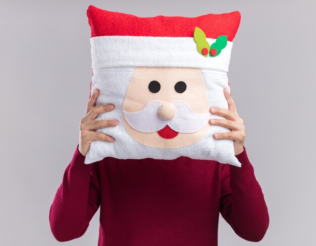Young guy wearing christmas hat with glasses covered face with christmas pillow isolated on white background