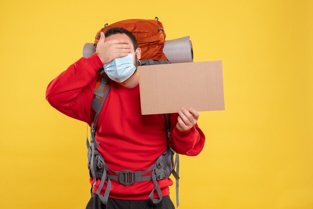 Young guy wearing medical mask with backpack and holding a sheet without writing putting hand on eyes on isolated yellow background