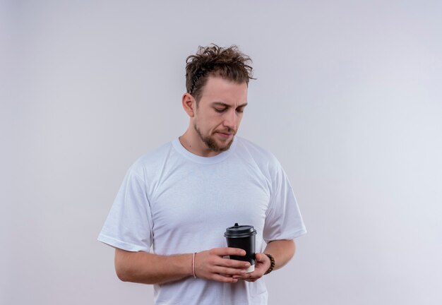 Молодой парень в белой футболке смотрит на чашку кофе на руке на изолированной белой стене