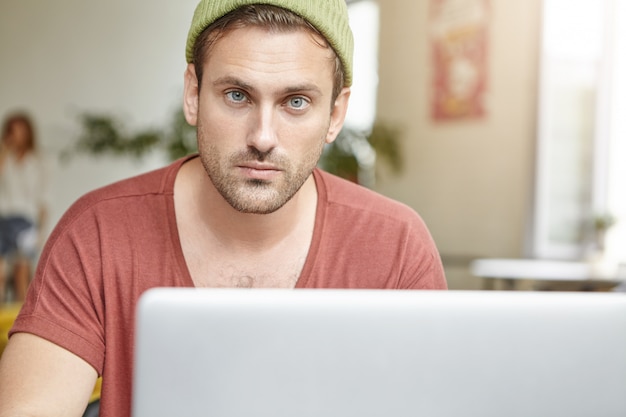 Giovane ragazzo con gli occhi azzurri e la barba guarda con sicurezza mentre si siede davanti al laptop aperto, controlla la posta elettronica o naviga sui social network online