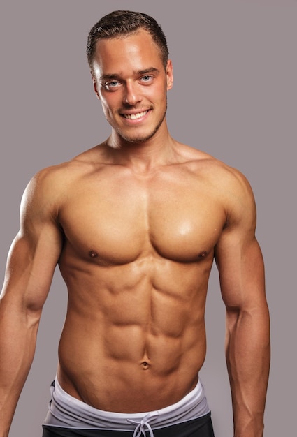Young guy with great body anatomy in black swim shorts possing in studio. Light grey background.