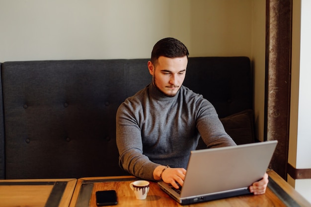 彼の携帯電話とコーヒーを持っている若い男。昼食時にシティカフェで若いファッションの男性エスプレッソコーヒーとラップトップコンピューターで作業