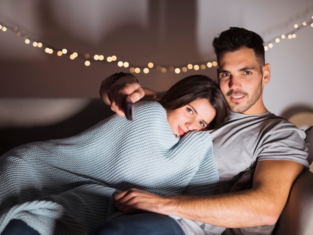 Young guy with TV remote hugging lady and lying on sofa