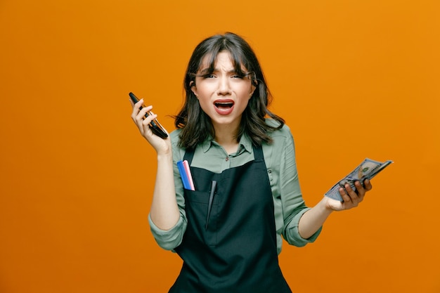 Young hairdresser woman wearing apron holding cash talking on mobile phone loooking confused and disappointed standing over orange background