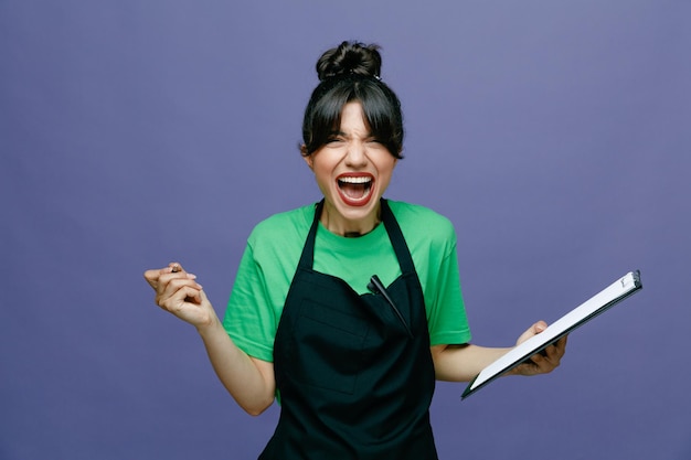 Young hairdresser woman wearing apron holding clipboard and pen shouting and yelling angry and frustrated standing over blue background