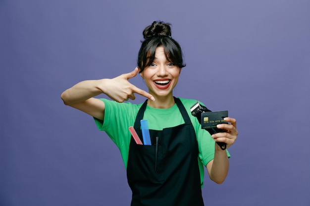 Молодая женщина-парикмахер в фартуке с электробритвой и кредитной картой, указывающая на нее указательным пальцем, смотрит в камеру, счастливая и позитивно улыбаясь, весело стоя на синем фоне