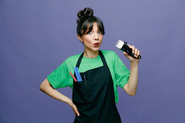 Young hairdresser woman wearing apron holding electric shaver looking at camera amazed and surprised standing over blue background