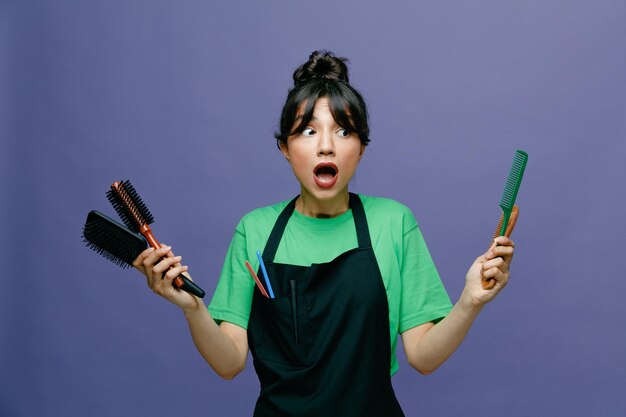 Young hairdresser woman wearing apron holding hair brushes looking amazed and surprised standing over blue background
