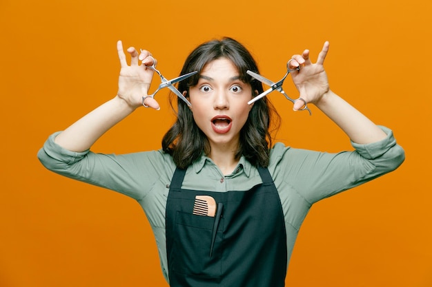 Young hairdresser woman wearing apron holding scissors looking at camera amazed and surprised standing over orange background