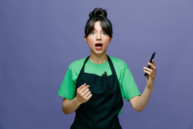 Young hairdresser woman wearing apron holding smartphone looking at camera amazed and surprised standing over blue background