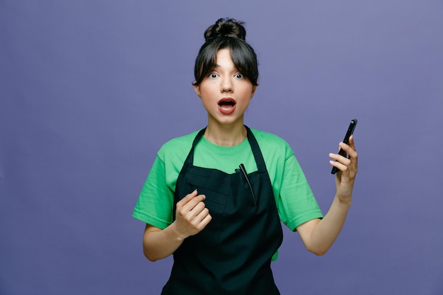 Молодая женщина-парикмахер в фартуке, держащая смартфон, смотрит в камеру, пораженная и удивленная стоя на синем фоне
