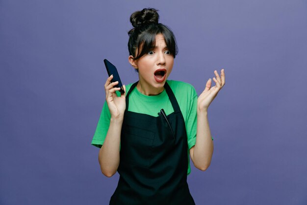 青い背景の上に立って心配して驚いたスマートフォンを持っているエプロンを着た若い美容師の女性