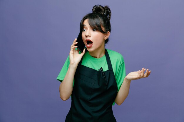 Young hairdresser woman wearing apron talking on mobile phone looking amazed and surprised standing over blue background