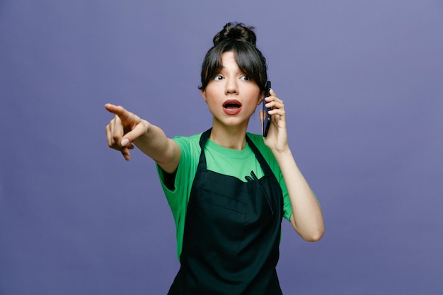 앞치마를 입은 젊은 미용사 여성이 파란색 배경 위에 서서 걱정스러운 것을 검지 손가락으로 가리키는 휴대전화로 통화하고 있다