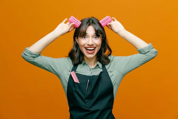 Young hairdresser woman wearing apron with comb holding hair curlers over her head like bunny ears looking at camera happy and positive smiling cheerfully standing over orange background
