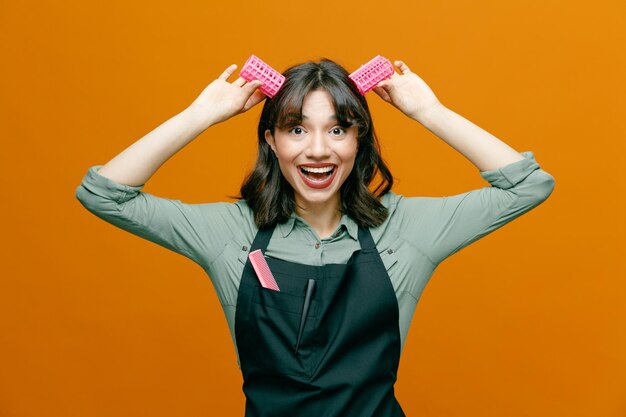 young hairdresser woman wearing apron with comb holding hair curlers over her head like bunny ears looking at camera happy and positive smiling cheerfully standing over orange background