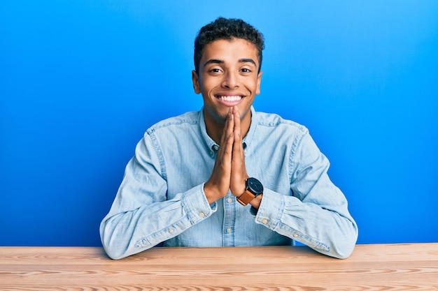 Бесплатное фото Молодой красивый афроамериканец в повседневной одежде сидит за столом и молится, сложив руки вместе, прося прощения, уверенно улыбаясь.