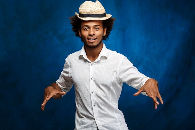 Young handsome african man in hat dancing over blue wall.