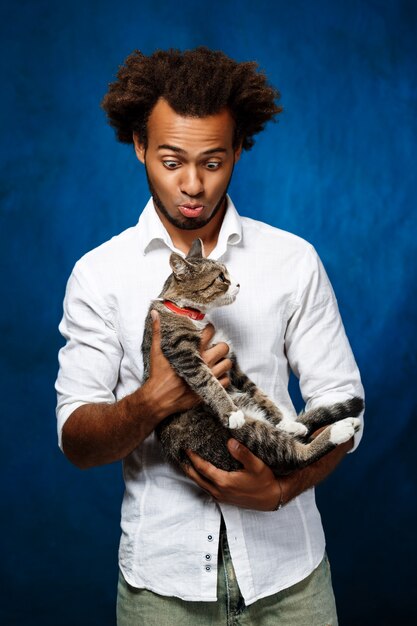 Young handsome african man holding cat over blue wall.