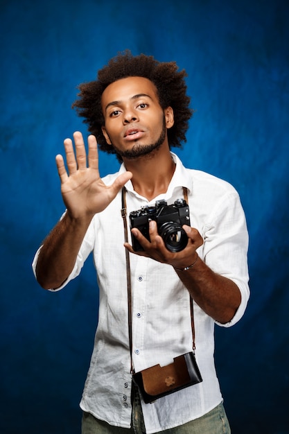 Молодой красивый африканский человек держа старую камеру над голубой стеной.