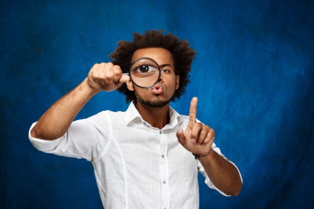 Young handsome african man posing with magnifier over blue wall.