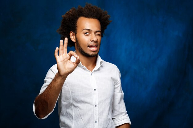 Young handsome african man showing okay, winking over blue wall.