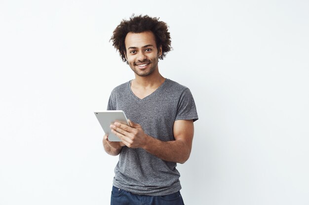 Young handsome african man smiling holding silver tablet and playing games or using a booking app against white wall.