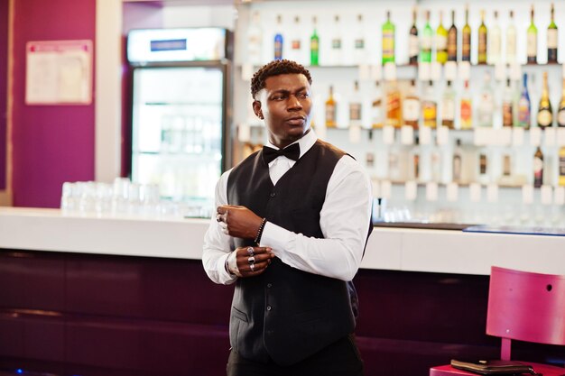 Young handsome african man wearing white shirt black vest and bow tie posed against bar counter at night club