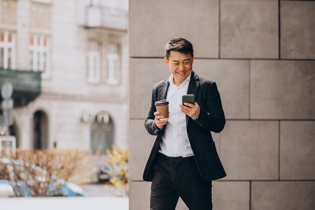 Молодой красивый азиатский деловой человек в черном костюме, используя телефон и пить кофе