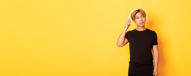 Young handsome asian guy with blond hair takeoff headphones to listen you standing yellow background