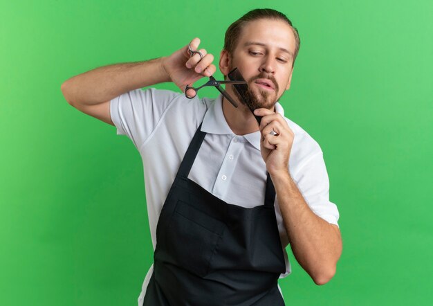 Young handsome barber wearing uniform combing and cutting his beard looking down isolated on green background