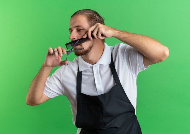 Young handsome barber wearing uniform combing and cutting his moustache looking at side isolated on green background