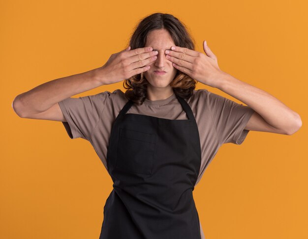 Young handsome barber wearing uniform covering eyes with hands isolated on orange wall