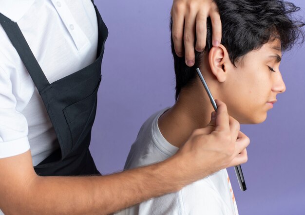 Young handsome barber wearing uniform doing haircut for young client isolated on purple background