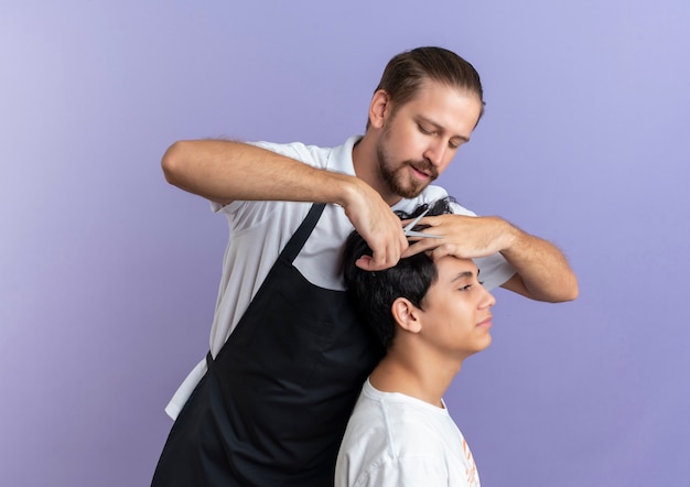 Young handsome barber wearing uniform doing haircut for young client isolated on purple background