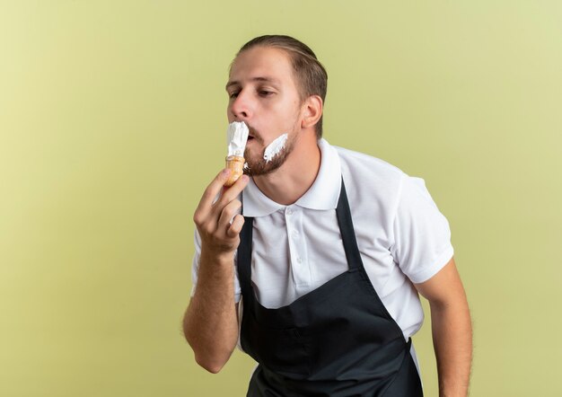 Young handsome barber wearing uniform holding shaving brush applying shaving cream on his own beard looking at side isolated on olive green background with copy space