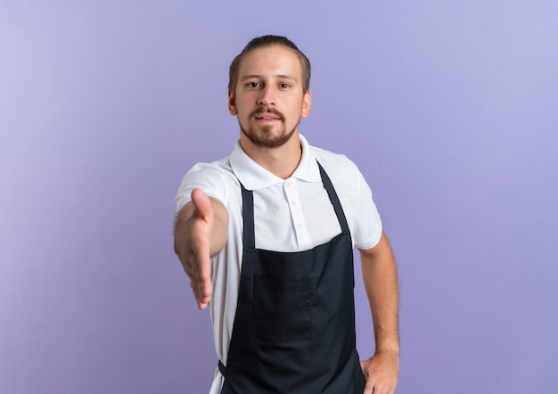 Young handsome barber wearing uniform putting hand on waist and stretching out another hand at camera doing hi gesture isolated on purple background with copy space