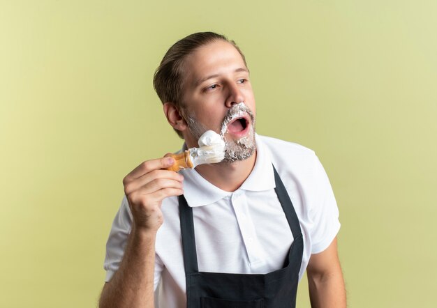 Young handsome barber wearing uniform putting shaving cream on his beard with shaving brush and looking at side isolated on olive green background