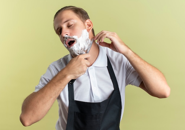 Young handsome barber wearing uniform shaving his own beard with straight razor with closed eyes isolated on olive green background